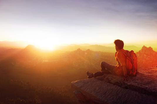 A guy is setting on the top of a mountain and watching a beautiful sunset. The light is so bright you almost can't see the landscape.