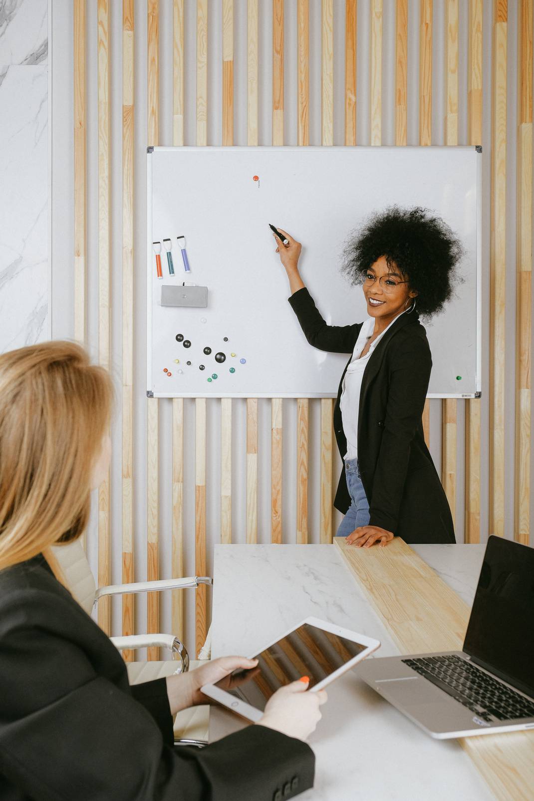 A black woman wearing glasses and a blazer is on a whiteboard while a blond woman in sitting looking to the whiteboard with a tablet on her hand